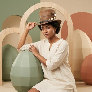 May include: A brown leather steampunk top hat with decorative metal accents, chain, and faux bullets. The hat is worn by a person wearing a cream-colored linen dress. The background features geometric shapes in shades of green, brown, and beige.