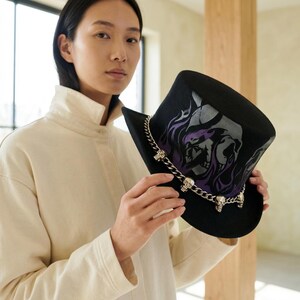 May include: Black top hat with a silver skull chain and a graphic design featuring a skull and purple flames. The hat is a stylish accessory.