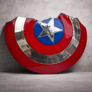 May include: A red, white, and blue shield with a silver trim and a star in the centre. The shield has a battle-worn appearance with a section missing from the top. The shield is leaning against a grey wall.