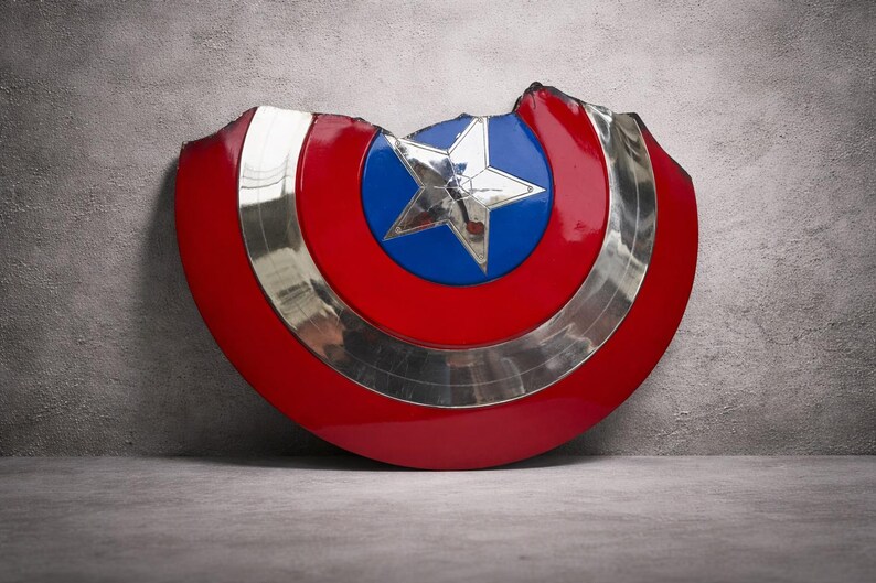 May include: A red, white, and blue shield with a silver trim and a star in the centre. The shield has a battle-worn appearance with a section missing from the top. The shield is leaning against a grey wall.