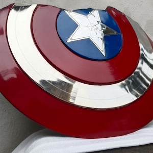 May include: A red, white, blue, and silver shield with a star emblem. The shield has a circular design with concentric rings of colour. The central emblem is a white star on a blue background. The shield is a prop or collectible.