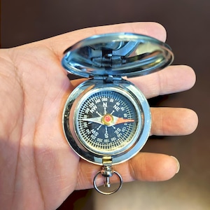 May include: A silver compass with an open lid, showing a black dial with white and red markings. The compass has a ring for attachment and is held in a hand.