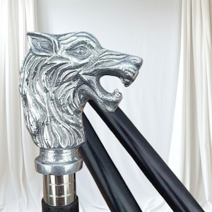 May include: A silver wolf head walking stick with a black shaft. The wolf head is intricately detailed, with visible fur and an open mouth. The stick has a metal collar and a dark wooden shaft.