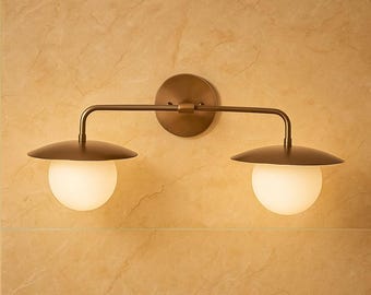 Puede incluir: Aplique de pared de color bronce con dos luces. Cada luz tiene un globo blanco redondo y una pantalla en forma de platillo de color bronce. La lámpara está montada sobre una base redonda de bronce.