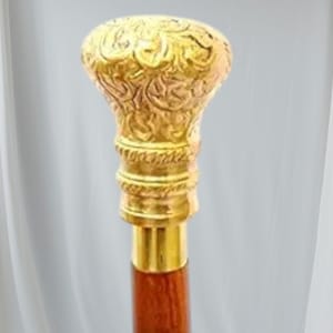 May include: A decorative walking cane with a gold-colored, ornate handle. The handle features detailed floral patterns and sits atop a brass collar and a dark brown wooden shaft. The cane is a classic accessory.