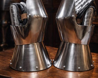 May include: A pair of polished steel gauntlets, featuring articulated finger plates and a flared cuff. The interior reveals black leather and brown accents. The gauntlets are displayed on a wooden surface, showcasing medieval armor.