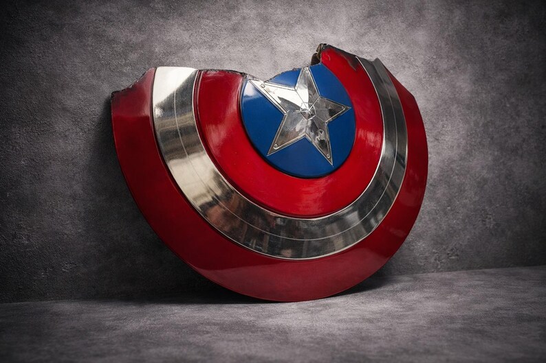 May include: A damaged Captain America shield. The shield has a red and silver design with a blue centre and a silver star. It is leaning against a grey textured surface, showcasing its iconic design.
