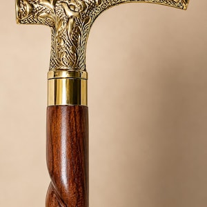 Op de afbeelding: Een wandelstok met een gepolijst, T-vormig handvat van messing, voorzien van gedetailleerde florale en ruitvormige reliëfpatronen. Het handvat is verbonden met een donkerbruine houten schacht met een opvallend spiraalvormig of gedraaid ontwerp.