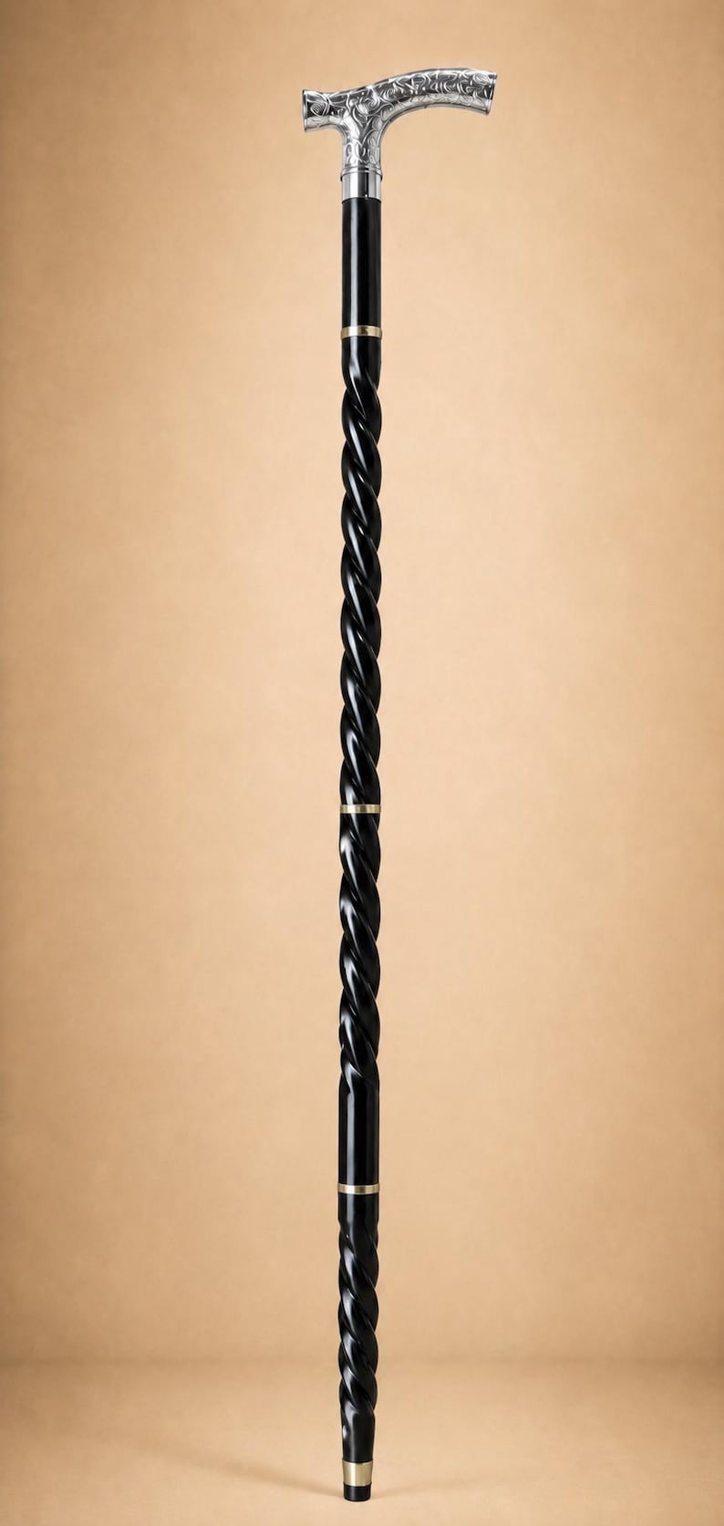 Op de afbeelding: Een wandelstok met een zwarte schacht in een gedraaid ontwerp en goudkleurige metalen ringen. Het handvat is gemaakt van gepolijst zilverkleurig metaal met een gedetailleerd reli&euml;fpatroon. De stok staat verticaal tegen een effen achtergrond.
