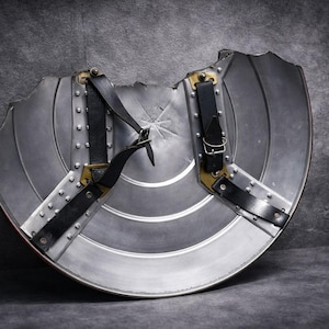 May include: A silver metal shield with a semi-circular shape, featuring horizontal ridges and a starburst design in the centre. It has black leather straps and brass-coloured hardware. The shield has a distressed appearance.