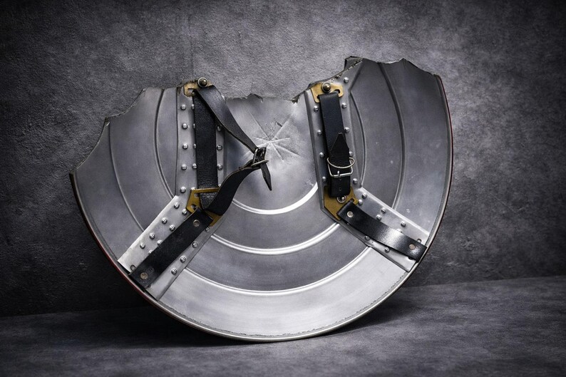May include: A silver metal shield with a semi-circular shape, featuring horizontal ridges and a starburst design in the centre. It has black leather straps and brass-coloured hardware. The shield has a distressed appearance.