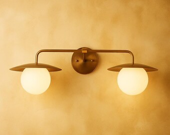 Puede incluir: Aplique de pared de latón con dos lámparas esféricas. Cada lámpara tiene un globo blanco y una pantalla circular. La lámpara está montada sobre una placa trasera redonda y tiene una barra horizontal.