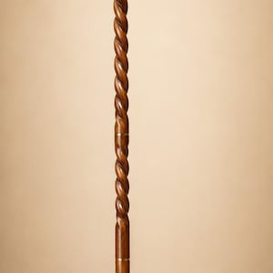 Op de afbeelding: Een wandelstok met een donkerbruine houten schacht met een doorlopend spiraalvormig ontwerp. Het handvat is een T-vormig goudkleurig metalen stuk met ingewikkelde bloemengravures. Goudkleurige metalen ringen accentueren de schacht, met een zwarte dop.