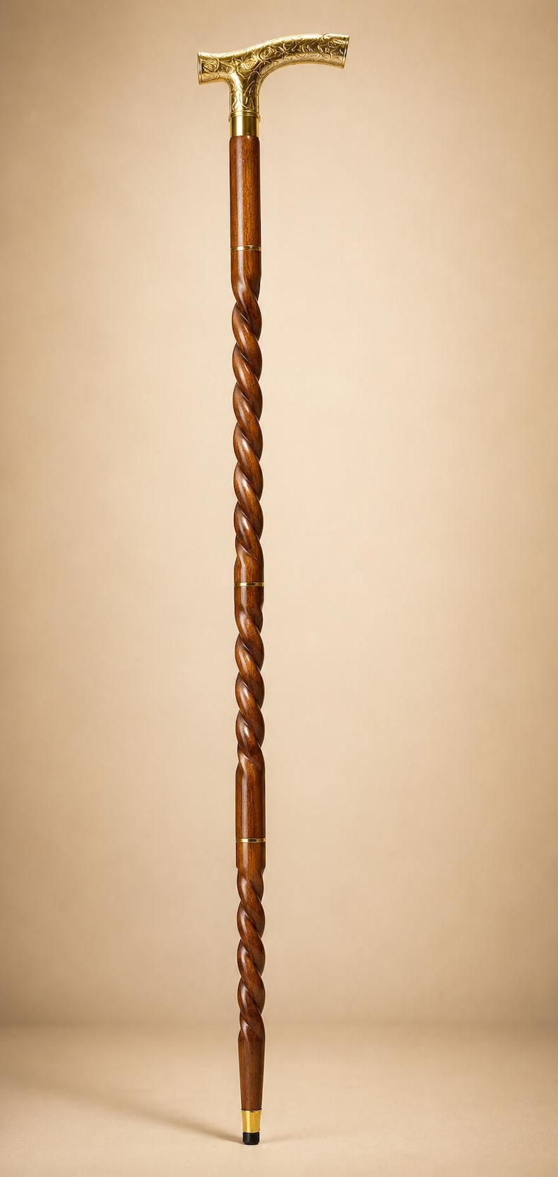 Op de afbeelding: Een wandelstok met een donkerbruine houten schacht met een doorlopend spiraalvormig ontwerp. Het handvat is een T-vormig goudkleurig metalen stuk met ingewikkelde bloemengravures. Goudkleurige metalen ringen accentueren de schacht, met een zwarte dop.