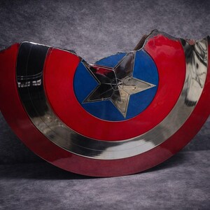 May include: A Captain America shield replica, with a red, white, and blue design. The shield has a silver metallic rim and a central blue circle with a silver star. The shield has a damaged section, giving it a battle-worn appearance.
