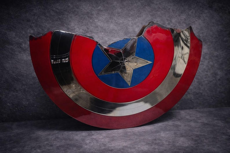 May include: A Captain America shield replica, with a red, white, and blue design. The shield has a silver metallic rim and a central blue circle with a silver star. The shield has a damaged section, giving it a battle-worn appearance.