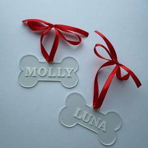 Op de afbeelding: Twee doorzichtige, botvormige huisdierlabels met rode strikken. Eén label leest "MOLLY" en de andere "LUNA" in een schreefloos lettertype. De labels hangen tegen een witte achtergrond.