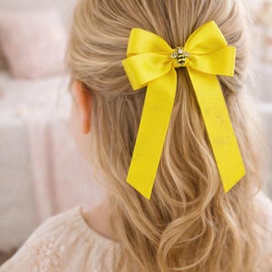 May include: A vibrant yellow hair bow, adorned with a bee-shaped embellishment, is styled in blonde hair. The bow is crafted from a broad ribbon, featuring long, flowing tails. The bee charm adds a playful element to the hair accessory.