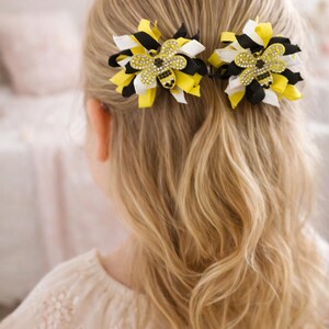 May include: Two flower-shaped hair clips with yellow, black, and white ribbons. Each clip features a rhinestone bee in the centre. The hair clips are in a young person's blonde hair.