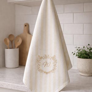 Può includere: Asciugamano da cucina a righe color crema e bianco con una ghirlanda ricamata in oro e la lettera "W". L'asciugamano è appeso a un gancio dorato. Un tagliere e utensili di legno sono sullo sfondo.