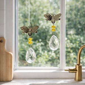 May include: Two decorative window hangings with bee and crystal drop designs. Each hanging features a metal bee, yellow beads, and a clear, teardrop-shaped crystal. The hangings are suspended from a silver chain, adding a touch of elegance to any space.