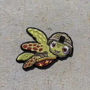 May include: An embroidered patch featuring a cartoon sea turtle wearing a camouflage military helmet. The turtle has light green skin, large brown eyes, and a shell with red and yellow patterns. The entire design is outlined in black thread.