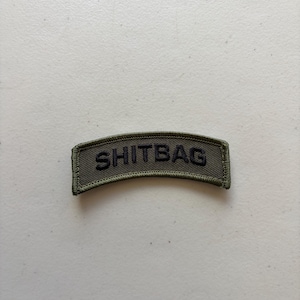 May include: An olive green, arched, rectangular embroidered patch with a dark border. The word SHITBAG is stitched in black, bold, capital letters across the center of the fabric. The patch features a textured, woven surface finish.