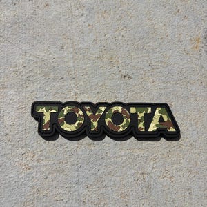 May include: A rectangular patch featuring the word TOYOTA in bold, block lettering. The letters have a camouflage pattern in shades of olive green, brown, tan, and cream, outlined by a thick, solid black border that follows the shape of the text.