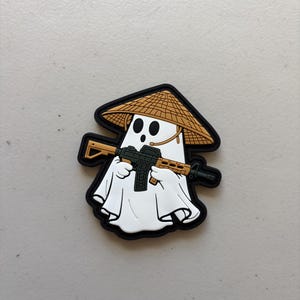 May include: A rubber morale patch featuring a white ghost character wearing a tan conical hat. The ghost is holding a black and tan rifle across its body. The entire design is outlined in a thick black border with a raised, textured finish.
