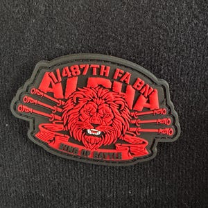 May include: A red and black patch featuring a lion's head with the text "ALPHA" and "1/487TH FA BN" above it. The text "KING OF BATTLE" is below the lion. The patch has a black border and a dark background.