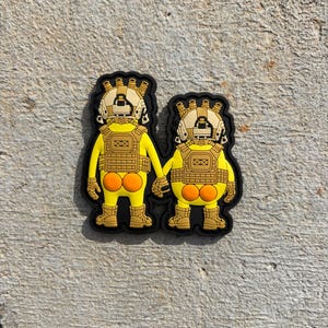 May include: A rubber morale patch featuring two yellow figures standing side-by-side and holding hands. Each figure wears a tan tactical helmet, a matching tactical vest, and tan boots, with exposed orange buttocks visible on their yellow bodies.