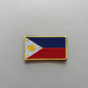 May include: A rectangular embroidered patch featuring the flag of the Philippines. The design includes a white triangle on the left with a yellow sun and three stars, alongside horizontal blue and red bands, all bordered by a yellow stitched edge.