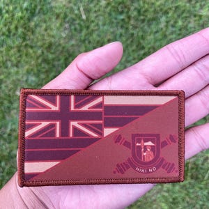 May include: A rectangular patch featuring the Hawaiian flag design. The patch is divided diagonally, showcasing the Union Jack and the state seal. The colors are primarily maroon, tan, and brown, with the text "HIKI NO" visible.