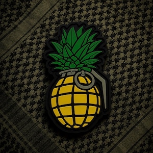 May include: A black patch featuring a yellow pineapple grenade design with green leaves. The grenade has a black outline and a silver ring. The background is a dark green and black patterned fabric.