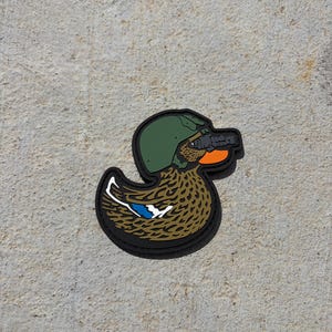 May include: A rubberized patch shaped like a mallard duck wearing a green military helmet and black night vision goggles. The duck has brown mottled feathers, an orange beak, and a blue and white wing patch, all outlined in black rubber.