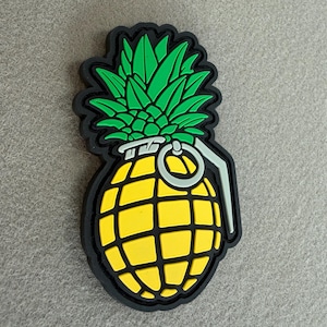 May include: A novelty item featuring a pineapple design, stylized as a hand grenade. The item has a yellow body with a grid pattern, green leaves, and a black outline. A silver ring and lever complete the design.