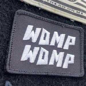 May include: A rectangular black patch with white embroidered text that reads "WDMP" stacked twice. The patch has a black border and is set against a dark, textured background. A partial view of a "TREAD ON ME" patch is visible.