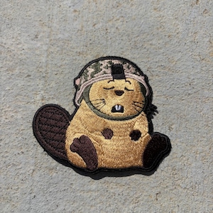 May include: An embroidered patch featuring a golden-brown beaver wearing a camouflage military-style helmet. The beaver has dark brown paws and a tail, with its eyes closed and two white front teeth visible in a resting facial expression.
