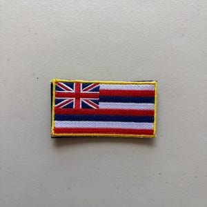 May include: A rectangular embroidered patch featuring the flag of Hawaii. The design includes a Union Jack in the upper left corner and eight horizontal stripes in alternating colors of white, red, and blue, all bordered by a bright yellow stitched edge.