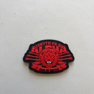 May include: A black and red tactical patch featuring a central lion head graphic. The text 1-487TH FA BN ALPHA is arched above the lion, and the text KING OF BATTLE is displayed on a banner below. Four artillery barrels extend from the sides.