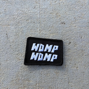 May include: A rectangular black embroidered patch with a stitched border. The patch features the text WDMP in white capital letters, repeated on two lines. The lettering has a bold, blocky font style with a slight slant to the characters.
