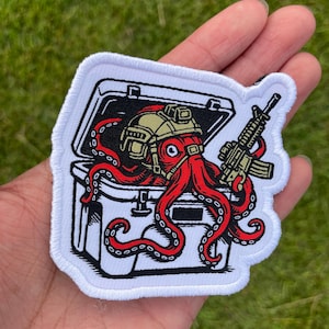 May include: Embroidered patch featuring a red octopus wearing a military helmet and holding a rifle. The octopus is emerging from a white container with a black interior. The patch has a white border and is likely for tactical gear.