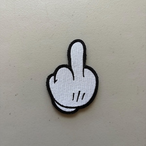 May include: A white embroidered patch featuring a cartoon-style hand with a black outline, displaying a middle finger gesture. The design includes three black vertical lines on the palm area to indicate detail, with a thick black border surrounding it.