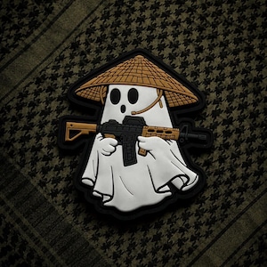 May include: A white ghost-shaped patch wearing a conical hat and holding a rifle. The patch has a black border and is set against a patterned background. The rifle has tan and black details.