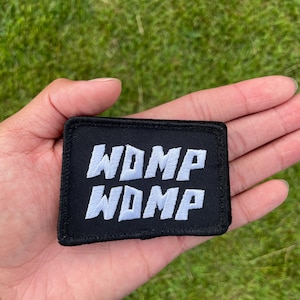 May include: A black rectangular patch with white embroidered text that reads "WDMP" stacked twice. The patch is held in a person's hand against a background of green grass.