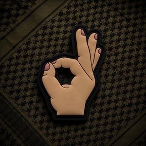 May include: A tan hand patch with a black border, forming the 'OK' gesture. The fingernails are painted pink. The patch is set against a dark green and black patterned background.