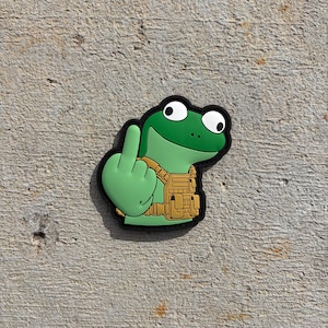 May include: A rubberized morale patch featuring a cartoon green frog with large white eyes, wearing a tan tactical vest, and gesturing with a middle finger. The design is outlined in black and has a smooth, molded surface with raised details.