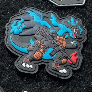 May include: A black and blue patch featuring a dragon-like creature holding a rifle. The creature has blue flames and accents, with brown and orange tactical gear. The patch has a black border and white claws.