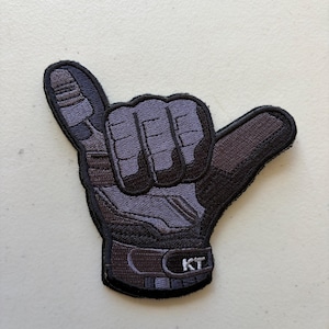 May include: An embroidered patch in the shape of a hand making a shaka sign. The design features a tactical glove in shades of dark gray and black, with the letters KT stitched in white on the wrist strap, finished with a black stitched border.