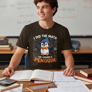 May include: A dark brown t-shirt with the text "I DID THE MATH: THE ANSWER IS PENGUIN" and a cartoon penguin wearing a blue hat and orange bow tie. The person is sitting at a desk with books and papers.